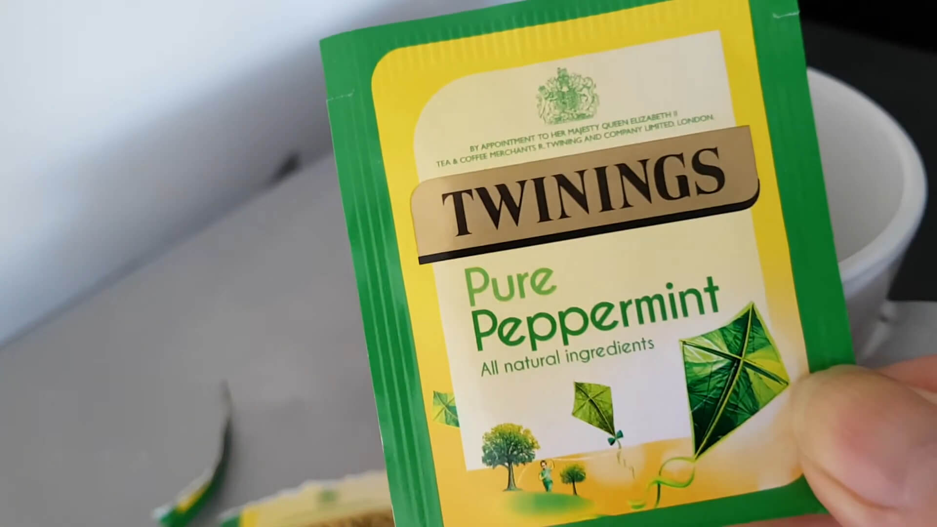 A hand holding a tea bag of peppermint, with a cup of brewed peppermint tea in the background