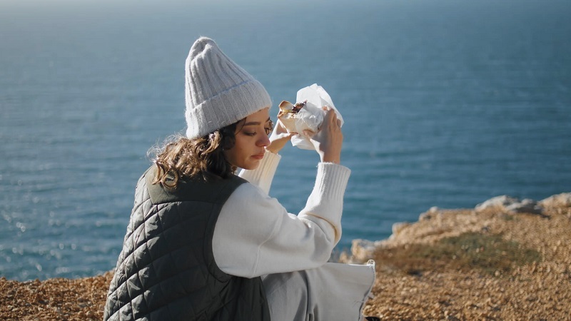 A woman in a white sweater and beanie sits by the ocean, unwrapping a sandwich for a snack while overlooking the serene blue waters