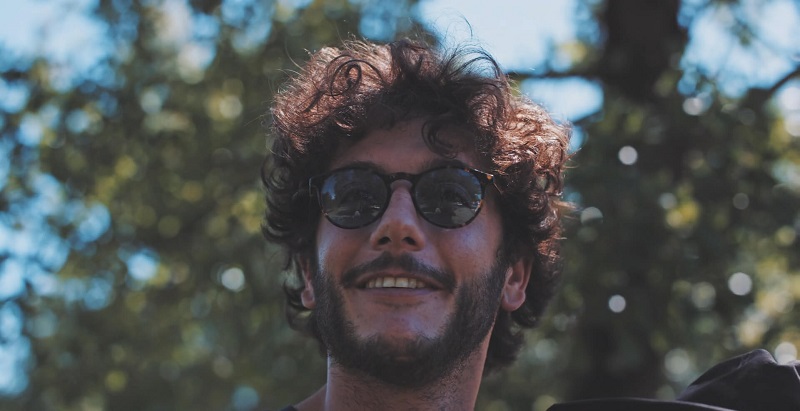 A man with curly hair and sunglasses smiles warmly in the sunshine, surrounded by the greenery of a park