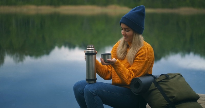 A young woman wearing a blue beanie and orange sweater pours herself a drink from a thermal flask while sitting by a peaceful lake