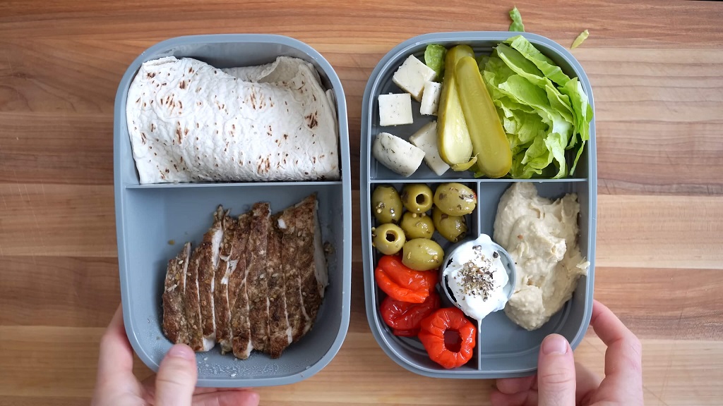 An insulated bento box with compartments containing sliced meat, stuffed peppers, olives, pickles, lettuce, and hummus