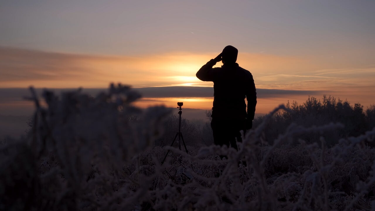 A photographer silhouetted against a golden sunrise in a frosty landscape, setting up a tripod