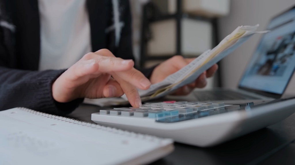 Close-up of a person using a calculator and reviewing documents while working at a desk, with a laptop and notepad in the background