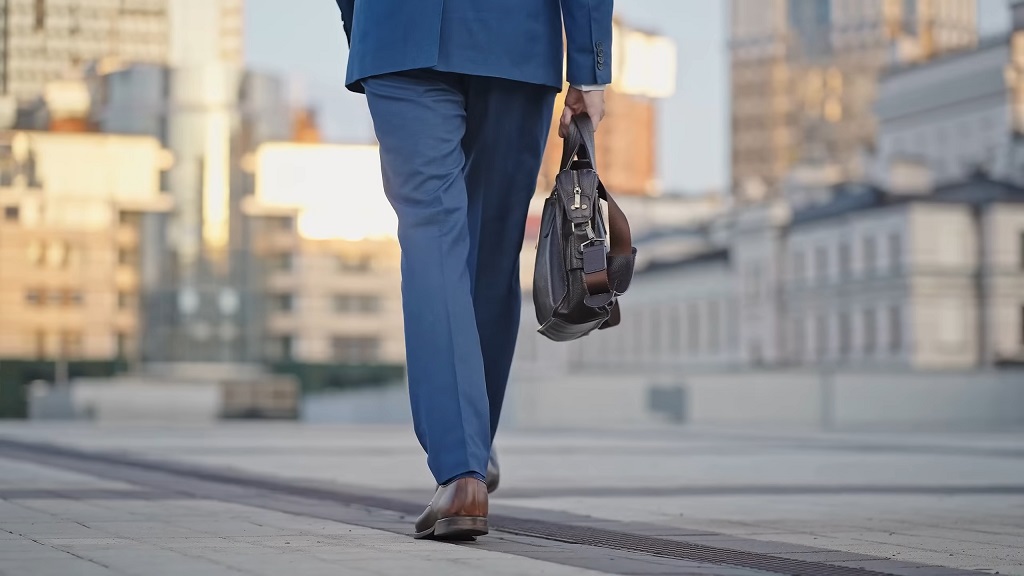 A person dressed in a blue suit walking on a paved urban street, holding a briefcase with modern office buildings in the background