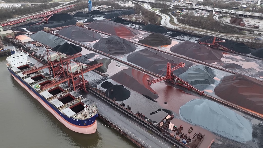 A busy port facility featuring a cargo ship being loaded with materials, and mounds of coal and other raw materials in storage areas