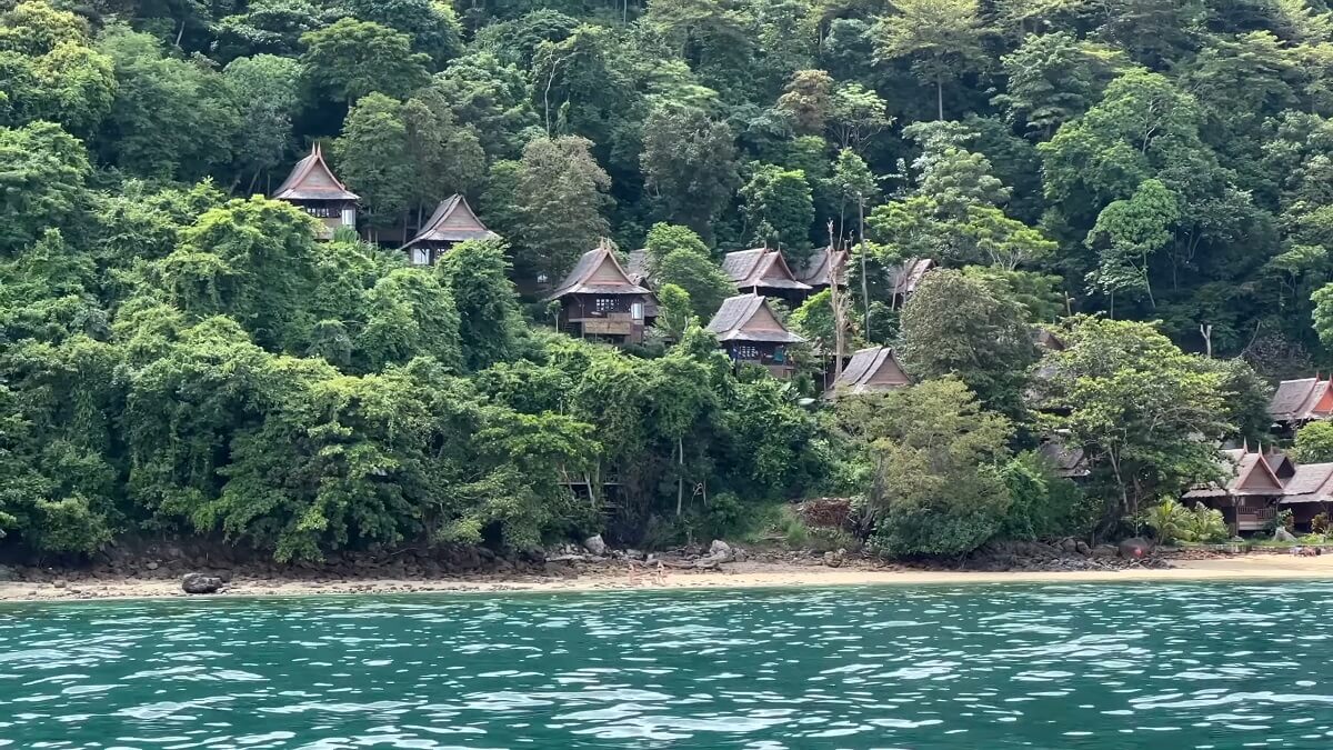 Traditional wooden huts with pointed roofs perched on a lush green hillside overlooking a turquoise sea in Thailand