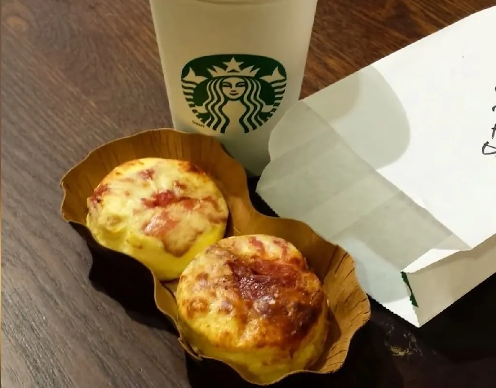 A pair of bacon and gruyere egg bites on a wooden table next to a Starbucks cup and a white bag