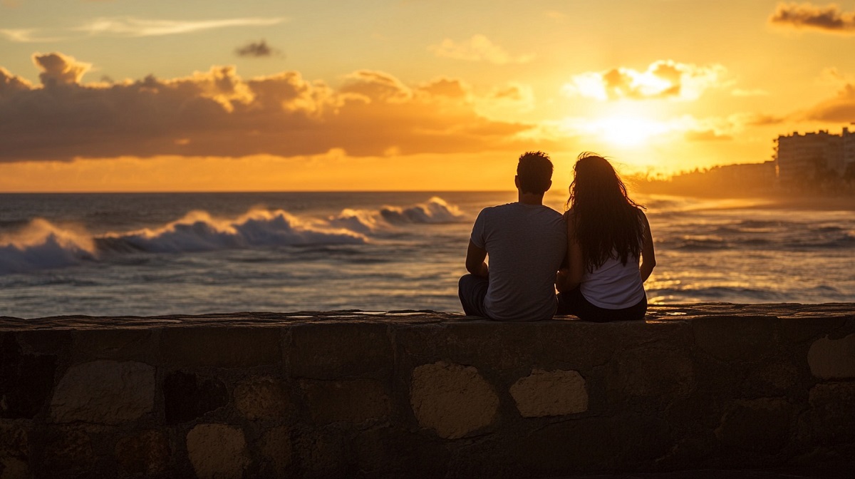 A couple sits on a stone wall overlooking the ocean waves as the sun sets in the background, creating a serene and intimate moment