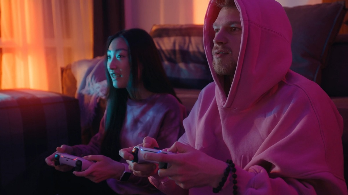 A young man and woman, both dressed in comfortable hoodies, sit on a couch, enjoying a video game together in a warmly lit room with a relaxed atmosphere