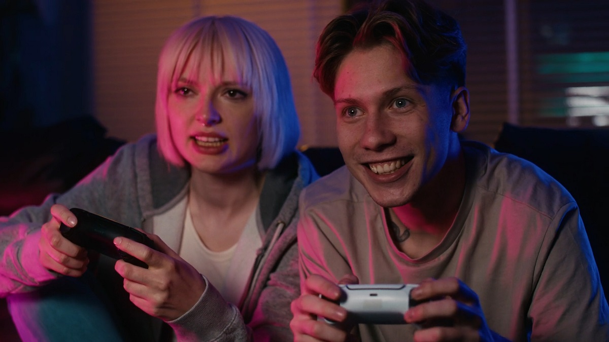  A young woman and a man excitedly playing video games together, holding controllers, illuminated by vibrant pink and blue lighting