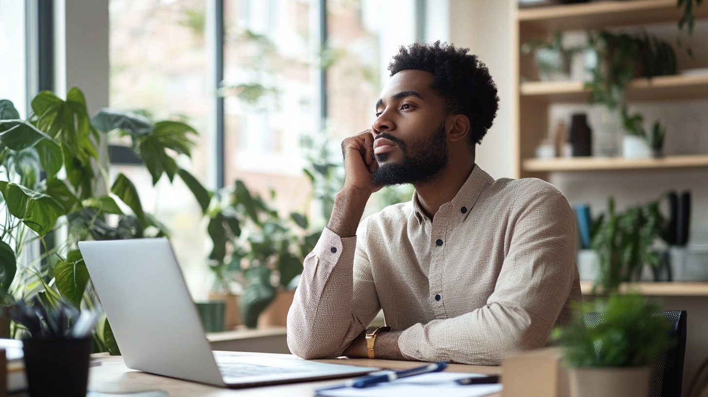 A young man sitting at a desk with a laptop, resting his chin on his hand, looking thoughtful in a bright, plant-filled office