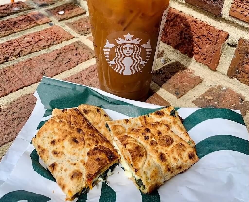 A Starbucks spinach, feta, and egg white wrap on branded packaging with an iced coffee against a brick background