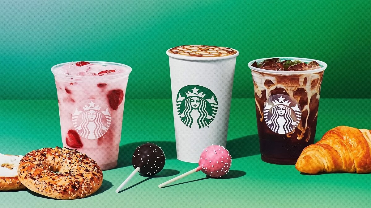 Three Starbucks beverages—a pink drink, a caramel macchiato, and an iced mocha—displayed with bagels, cake pops, and a croissant